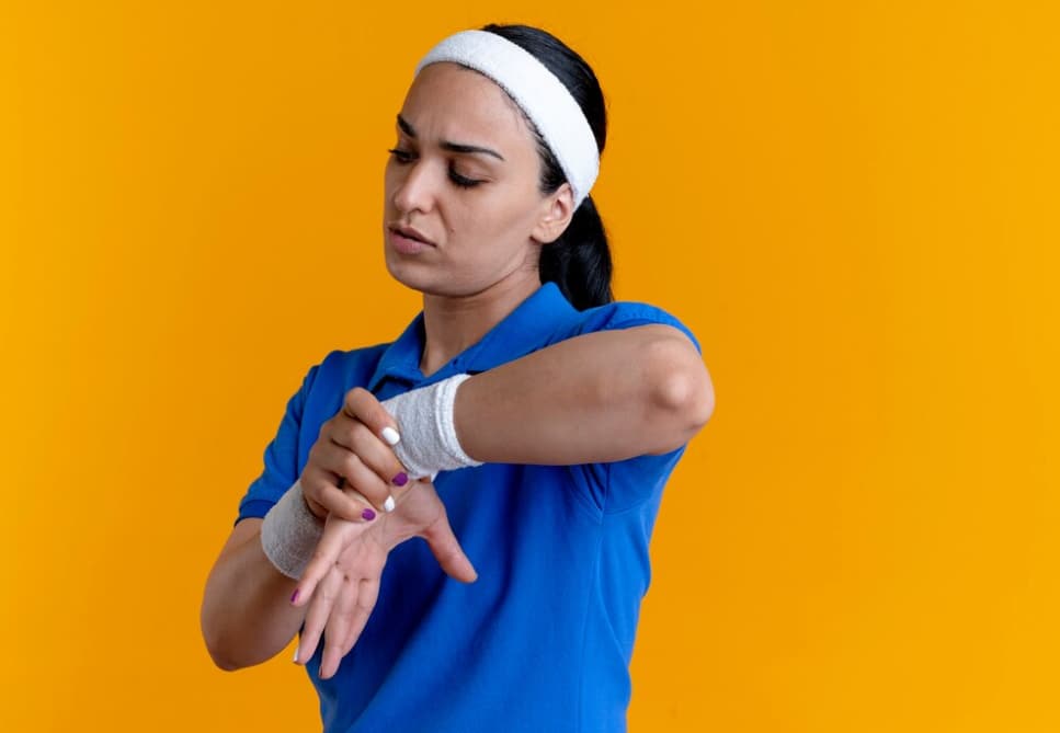 woman with a white headband and blue polo t-shirt holds her wrist in front of an orange wall